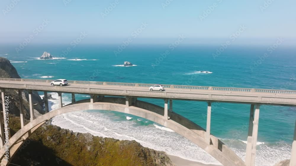 Cars driving slow by scenic arch architecture bridge with blue ocean background. Cinematic ocean waves under blue sky on sunny summer day. West coast road trip, eco nature travel 4K drone flight USA Cars driving slow by scenic arch architecture bridge with blue ocean background. Cinematic ocean waves under blue sky on sunny summer day. West coast road trip, eco nature travel 4K drone flight USA