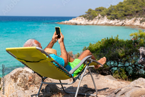 Mature man on holiday relax in the sun on the deck chair holding his phone chatting with friends.Vacation and technology.