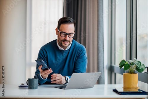 Professional messaging on smart phone while working on laptop at office desk.