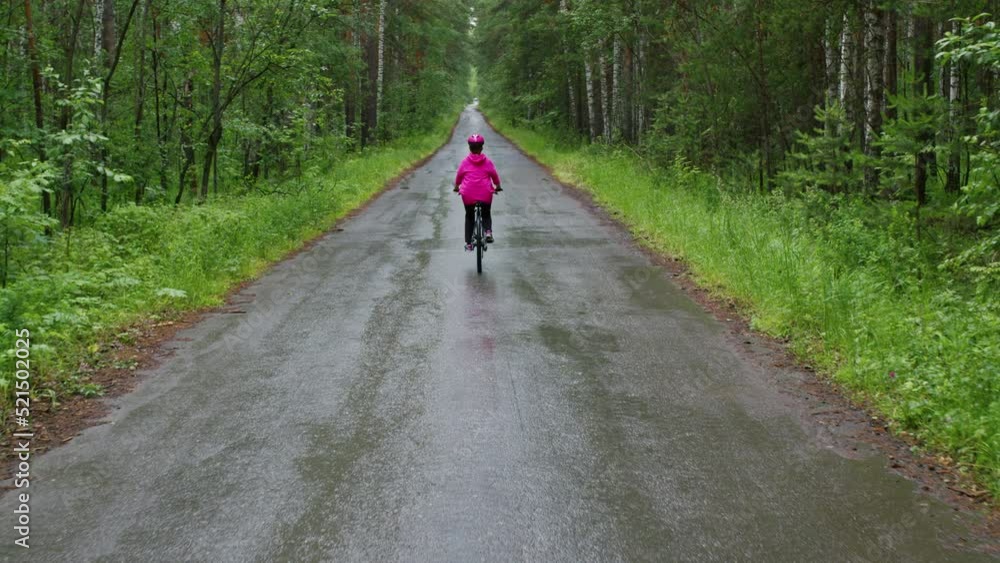 Woman in pink helmet, tracksuit rides bicycle on road in forest. Back view. Motion of female cyclist along highway surrounded by trees. Cycling amateur participates in marathon in wooded area Woman in pink helmet, tracksuit rides bicycle on road in forest. Back view. Motion of female cyclist along highway surrounded by trees. Cycling amateur participates in marathon in wooded area