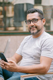handsome young man with glasses looking at his smartphone sitting in cafe outside