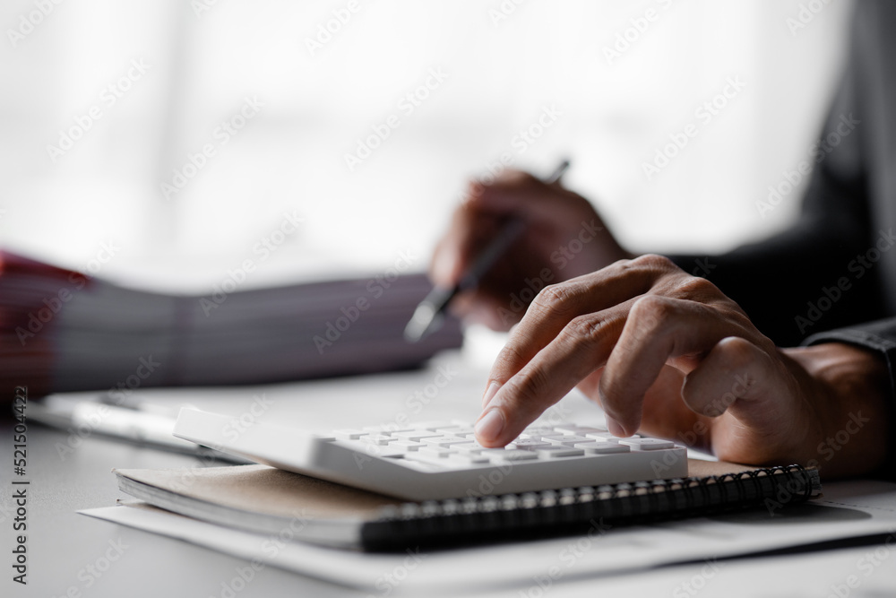 Businessman is using a calculator to calculate company financial figures from earnings papers, a businessman sitting in his office where the company financial chart is placed. Banner with copy space. Businessman is using a calculator to calculate company financial figures from earnings papers, a businessman sitting in his office where the company financial chart is placed. Banner with copy space.