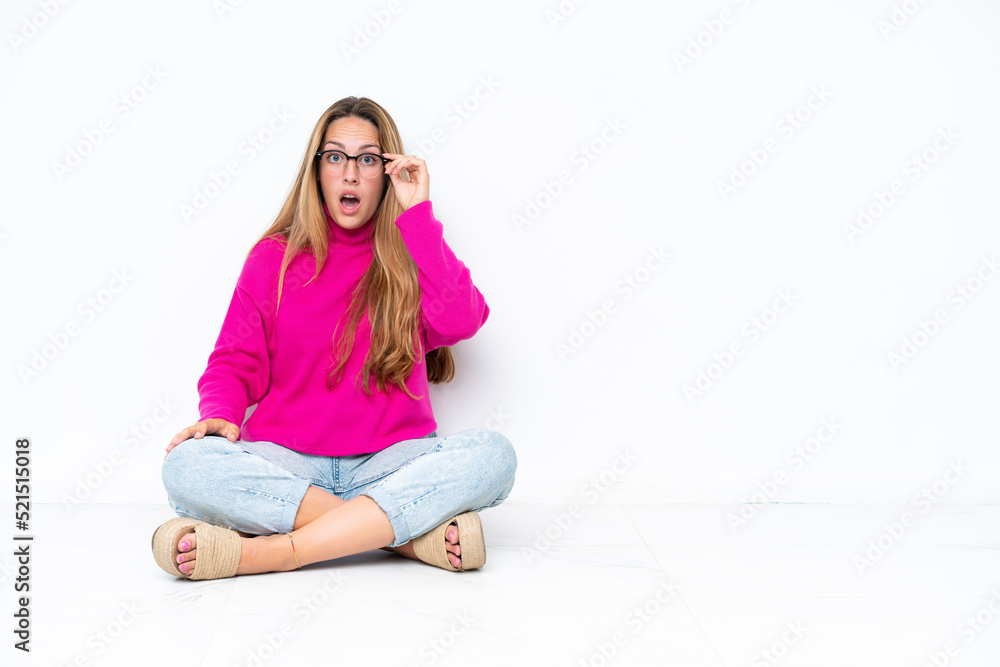 Young caucasian woman sitting on the floor isolated on white background with glasses and surprised Young caucasian woman sitting on the floor isolated on white background with glasses and surprised