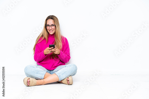 Young caucasian woman sitting on the floor isolated on white background sending a message with the mobile