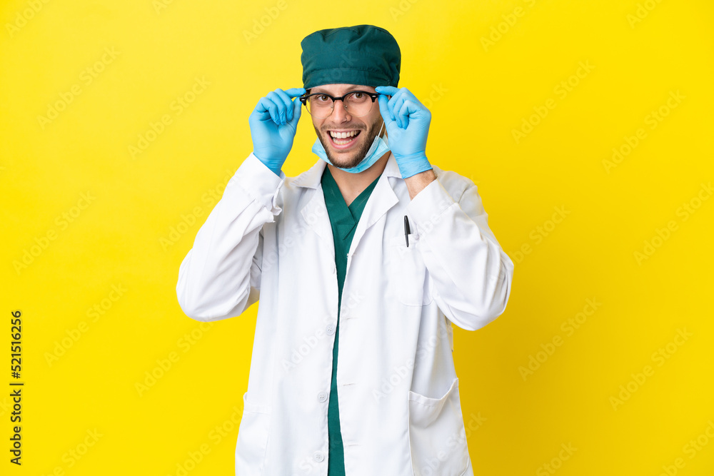 Surgeon blonde man in green uniform isolated on yellow background with glasses and surprised Surgeon blonde man in green uniform isolated on yellow background with glasses and surprised