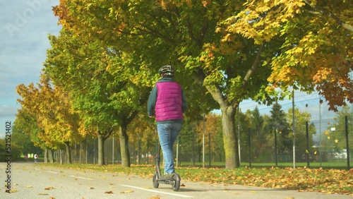 Rear view of an elderly woman driving an electric scooter next to autumn trees. Senior lady using modern end eco-friendly way of commuting. Beautiful autumn day to ride with e-scooter around the town. photo
