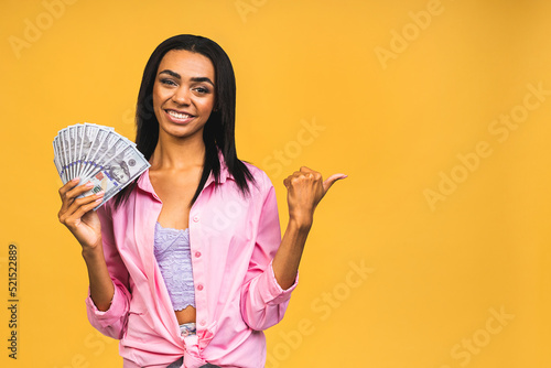 Happy winner. Portrait of african american successful woman 20s with afro hairstyle holding lots of money dollar banknotes isolated over yellow background. Pointing finger away.