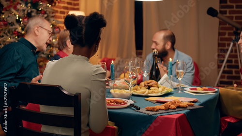 Multiethnic people gathered in living room to celebrate winter holiday while giving each other gifts. Festive family members sitting at Christmas dinner table while exchanging presents at home.
