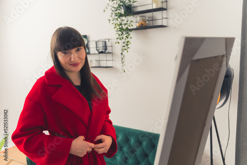 Charming young brunette woman putting on a red coat in front of the mirror in her apartment. High-quality photo