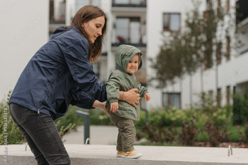 A mom is teaching her female toddler how to do steps in the street at noon.