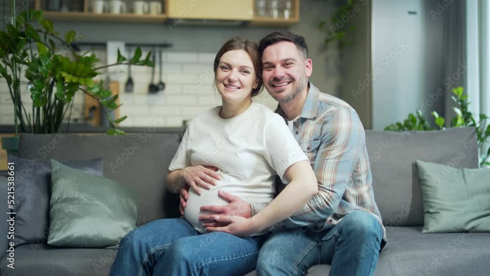 portrait happy family during pregnancy at home. Young happy family Caucasian couple husband and pregnant wife with belly sitting on sofa in living room and looking at camera smiling. Indoor. Happiness portrait happy family during pregnancy at home. Young happy family Caucasian couple husband and pregnant wife with belly sitting on sofa in living room and looking at camera smiling. Indoor. Happiness
