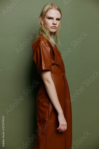 Self-confident fashionable blonde young model looking over her shoulder at camera dress in brown leather trench coat posing isolated on over olive green studio wall background. Stylish offer © SHOTPRIME STUDIO