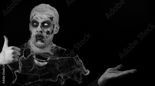 Zombie man with makeup with fake wounds scars showing thumbs up and pointing empty place, advertising area for commercial text logo, copy-space for goods promotion. Sinister Halloween ugly dead guy