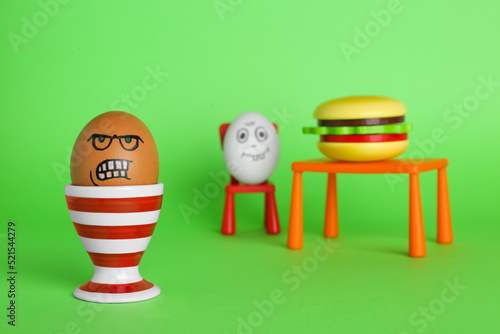 Angry egg and happy one with burger at table on green background