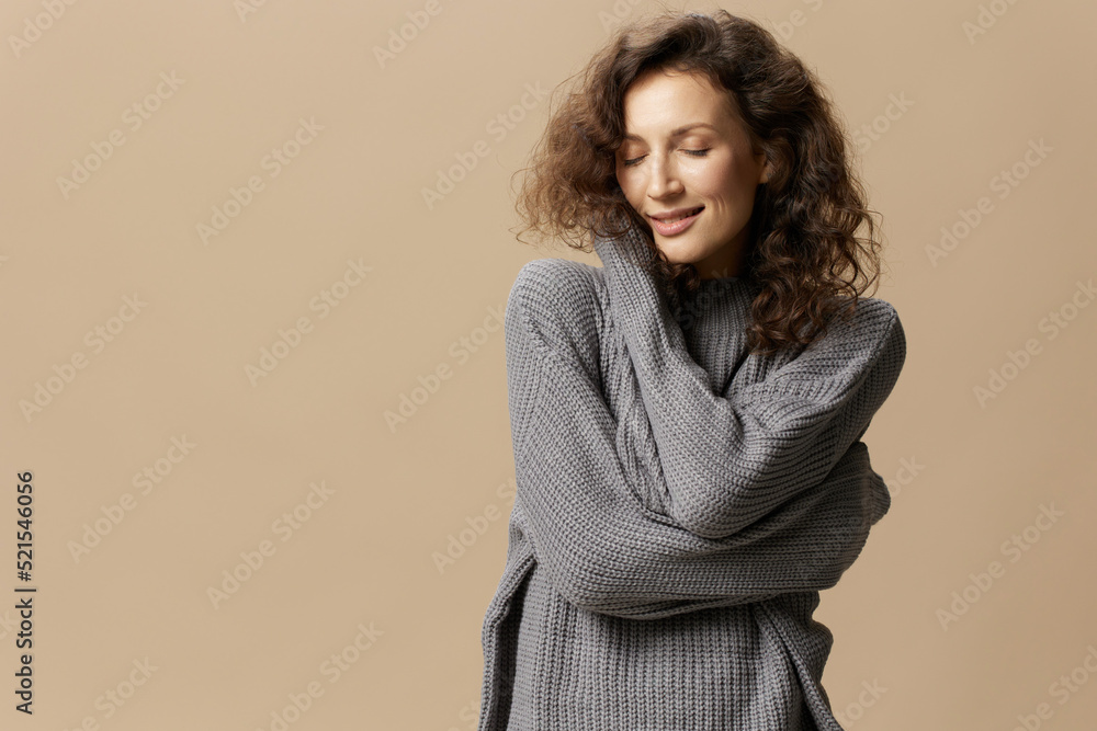 Enjoyed happy curly beautiful female in gray warm casual sweater hugs herself closing eyes posing isolated on over beige pastel background. Fashion Sale offer. People emotions concept. Copy space Enjoyed happy curly beautiful female in gray warm casual sweater hugs herself closing eyes posing isolated on over beige pastel background. Fashion Sale offer. People emotions concept. Copy space