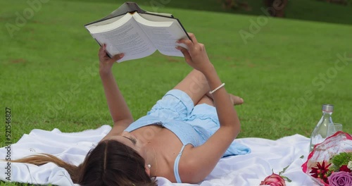 Young latina in a blue dress reading a book while laying in the park at a picnic setting photo