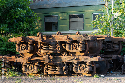 Rusting wagon bogies and old railway equipment from electric locomotives in Tbilisi, Georgia in stock on street on sunny day. Decommissioned soviet engineering put on ground near lush green trees