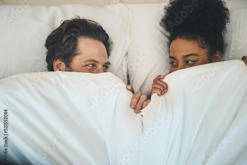 Intimate, loving and affectionate couple or lovers in bed after a passionate night or sensual sex. Playful interracial partners having fun or hiding under blanket on a romantic honeymoon.