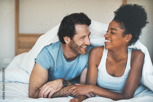 Happy, carefree and laughing couple having fun lying in bed together. Interracial husband and wife bonding and showing affection while talking. Smiling lovers enjoying time indoors being playful photo