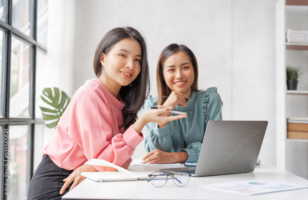 Two Young charming Asian businesswoman discussing and analyze planning and financial statistics when sitting at desk in workplace. Looking camera. Two Young charming Asian businesswoman discussing and analyze planning and financial statistics when sitting at desk in workplace. Looking camera.