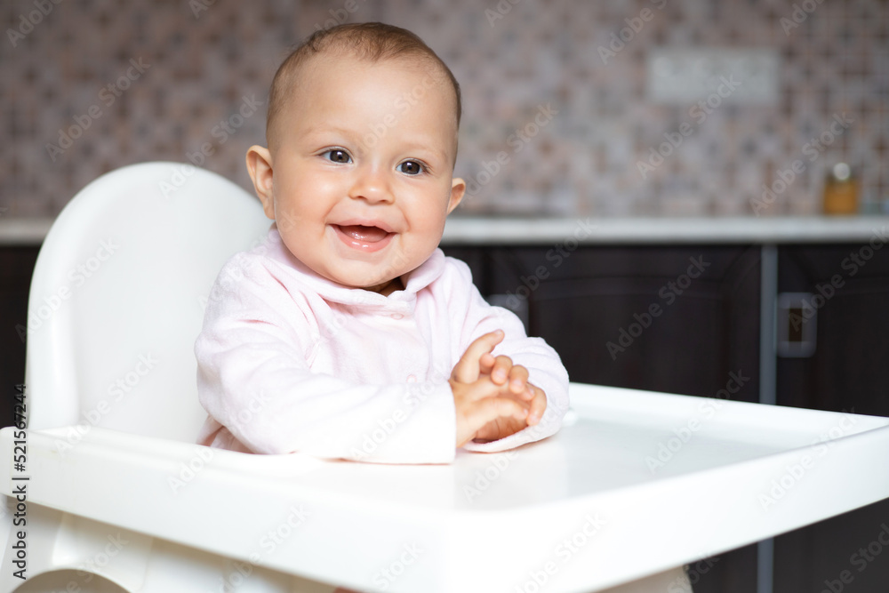 Cute Baby in pink clothes sits in a highchair in the kitchen. Space for text. Lifestyle Cute Baby in pink clothes sits in a highchair in the kitchen. Space for text. Lifestyle