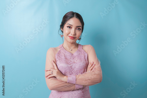 Portrait of a confident smiling girl standing with arms folded and looking at camera isolated over blue background, wearing modern kebaya dress