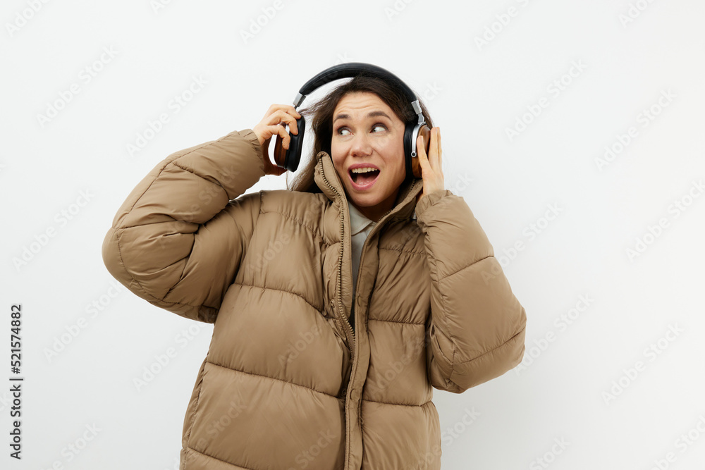 a beautiful woman in a stylish warm down jacket stands on a light background putting on headphones to listen to music a beautiful woman in a stylish warm down jacket stands on a light background putting on headphones to listen to music