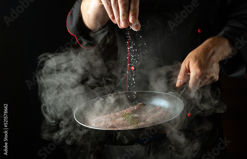 Fototapeta Naklejka Na Ścianę i Meble -  The chef adds salt to a hot frying pan with sausages. Menu or recipe concept for hotel on black background