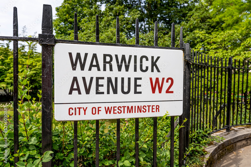 Metal and white street name sign on a fence pointing Warwick Avenue, in London, City of Westminster, England, United Kingdom