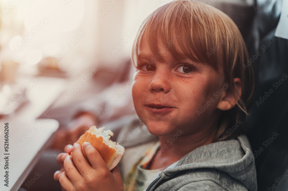 little candid kid boy five years old eats burger or sandwich food sitting in airplane seat on flight traveling from airport. children take a bite. child in air plane eating lunch or dinner meal. flare little candid kid boy five years old eats burger or sandwich food sitting in airplane seat on flight traveling from airport. children take a bite. child in air plane eating lunch or dinner meal. flare