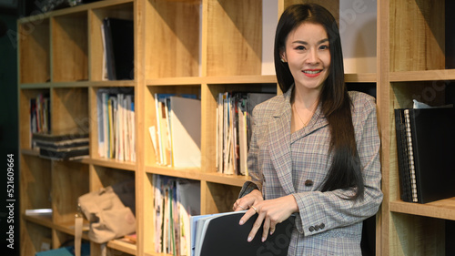 Portrait of millennial working woman in business suit standing near bookshelves in her personal office and smiling at camera