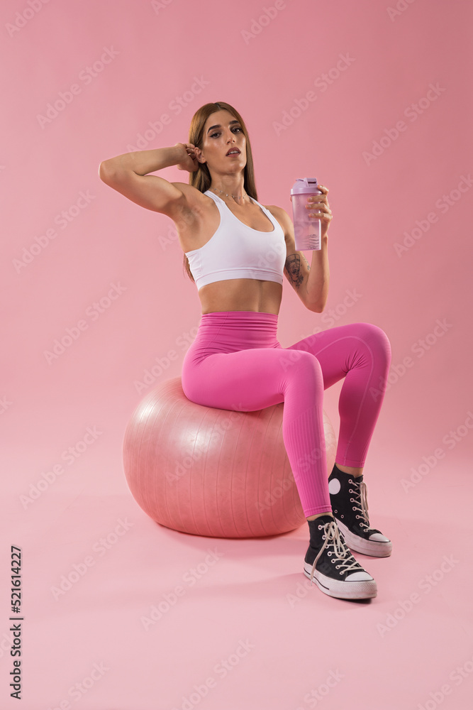 Fitness girl in pink sportswear posing on top of a yoga ball while holding a bottle on a pink background. Sport concept. Fitness girl in pink sportswear posing on top of a yoga ball while holding a bottle on a pink background. Sport concept.