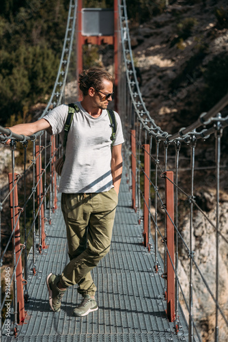 A man with a backpack on a suspension bridge against the backdrop of a mountain landscape with trees. Hiking trail pass through the hanging bridge. Travel and exploration concept photo