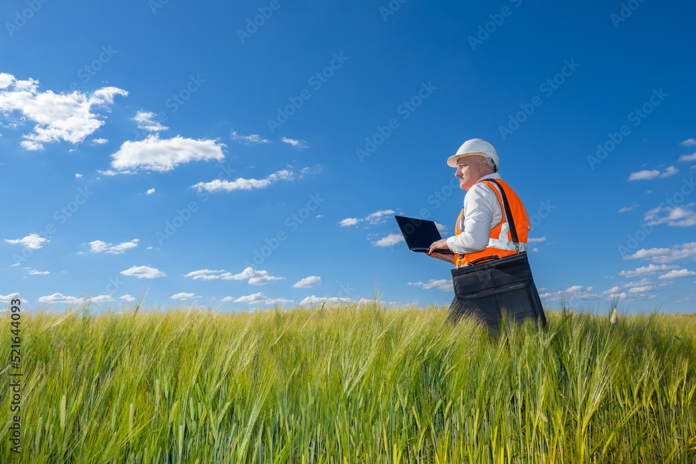 Builder walks across field with grass. Civil engineer with laptop. Future Construction site. Man in protective helmet inspects field. Engineer in reflective vest. Engineer on background sky and grass