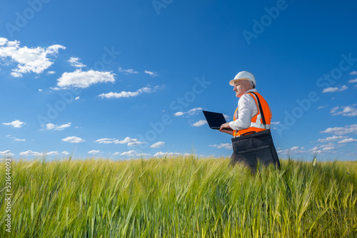 Builder walks across field with grass. Civil engineer with laptop. Future Construction site. Man in protective helmet inspects field. Engineer in reflective vest. Engineer on background sky and grass