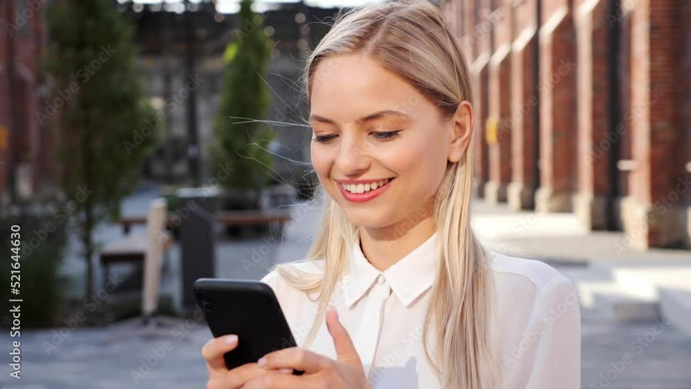 Caucasian smiling successful businesswoman in white shirt standing in urban street, taping on mobile phone communicates in social networks scrolling smartphone outdoor. Female freelancer relaxing. Caucasian smiling successful businesswoman in white shirt standing in urban street, taping on mobile phone communicates in social networks scrolling smartphone outdoor. Female freelancer relaxing.