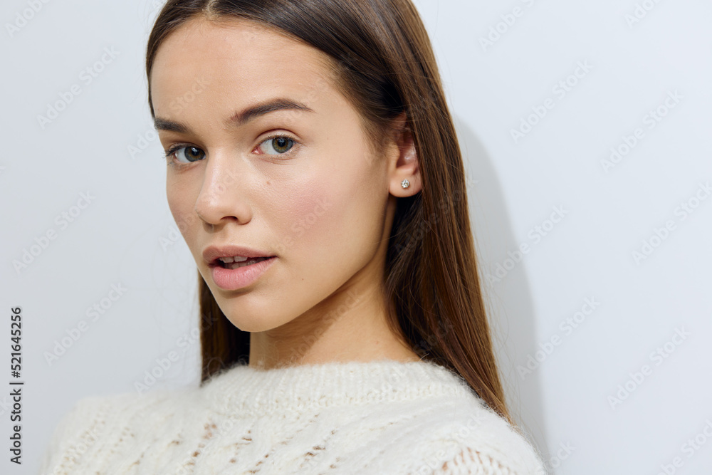 close horizontal portrait of a beautiful, well-groomed woman standing on a light background in a white, stylish, warm sweater, looking playfully at the camera with her hair tied behind her ears close horizontal portrait of a beautiful, well-groomed woman standing on a light background in a white, stylish, warm sweater, looking playfully at the camera with her hair tied behind her ears