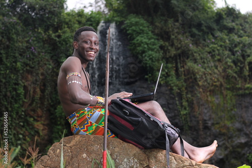 Education in Africa,online classes in Africa: A young African Masai Moran warrior sits on a rock beside a waterfall studying using a laptop 