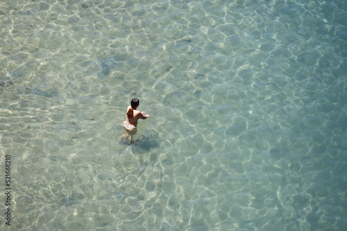 Aerial view to azure sea and woman in bikini go to swim in transparent water. Girl swimmer, beach vacation in summer