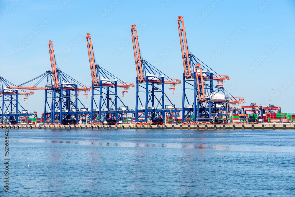 Tall gantry cranes on a commercial dock with stacks of colourful containers in background on a sunny summer day 