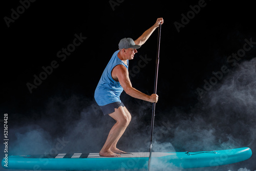 A man in a cap on a sub board with a paddle on a black background in the fog.