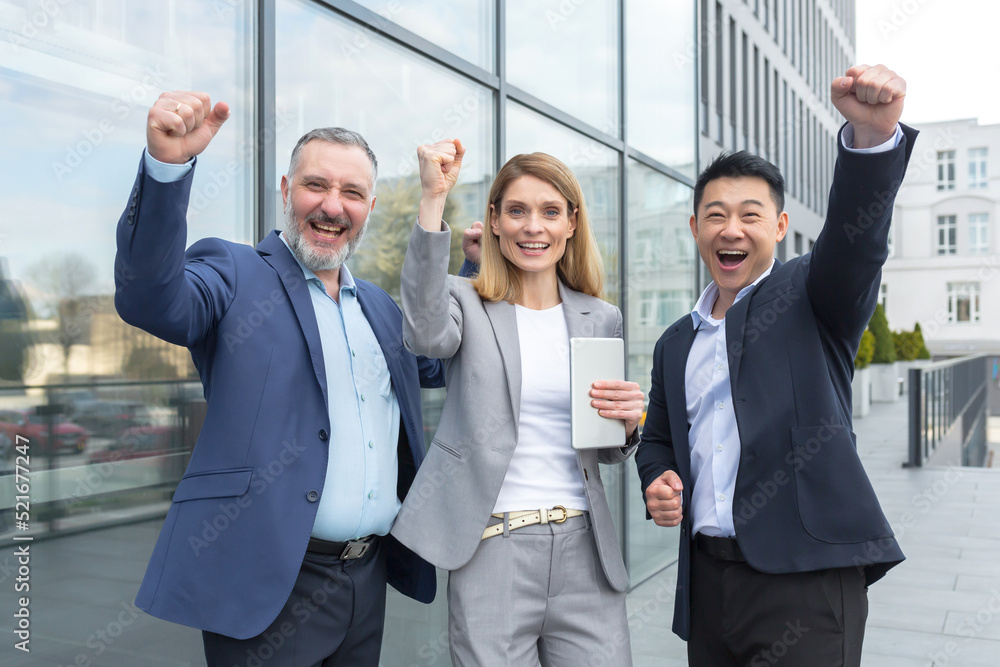 Successful diverse business team, business people looking at camera and rejoicing in success and victory, man and woman holding hands up outside office Successful diverse business team, business people looking at camera and rejoicing in success and victory, man and woman holding hands up outside office