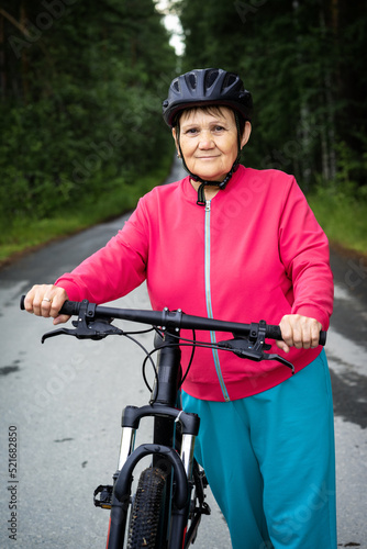 Elderly woman in helmet stands next to bicycle on forest road. Senior lady doing cycling in wooded place. Healthy, sportive, active lifestyle in old age