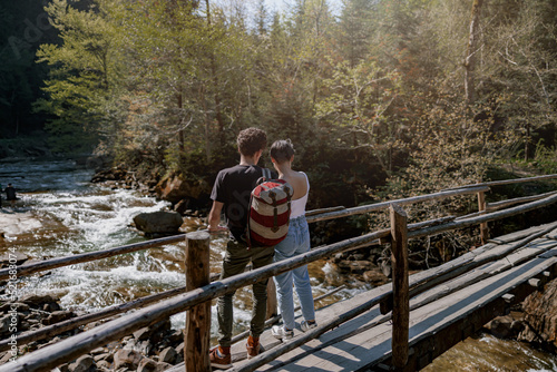 Back view on couple of young hikers standing on wooden bridge over fast stream and hugging.