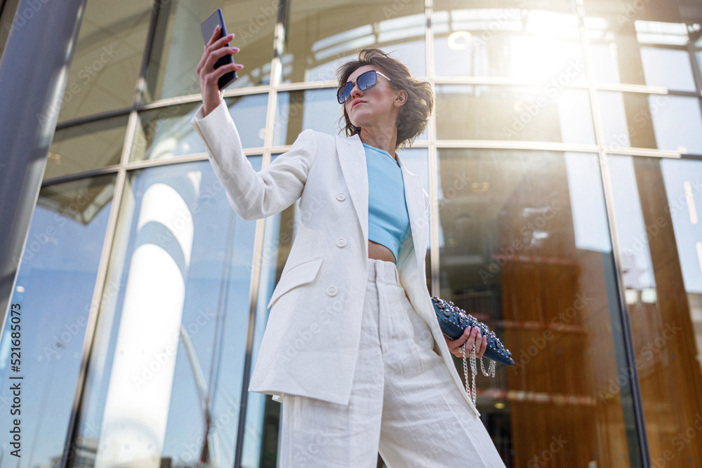 Pretty woman in trendy outfit making selfie on background of modern building
