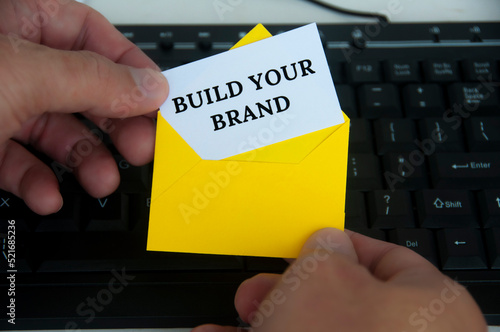 Hand holding with notepad in yellow envelope with text - Build your brand. With keyboard monitor background.