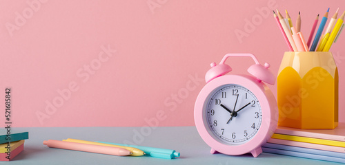 Back to school concept. Photo of school accessories on blue table pencil holder alarm clock stack of notebooks and pens on pastel pink wall background