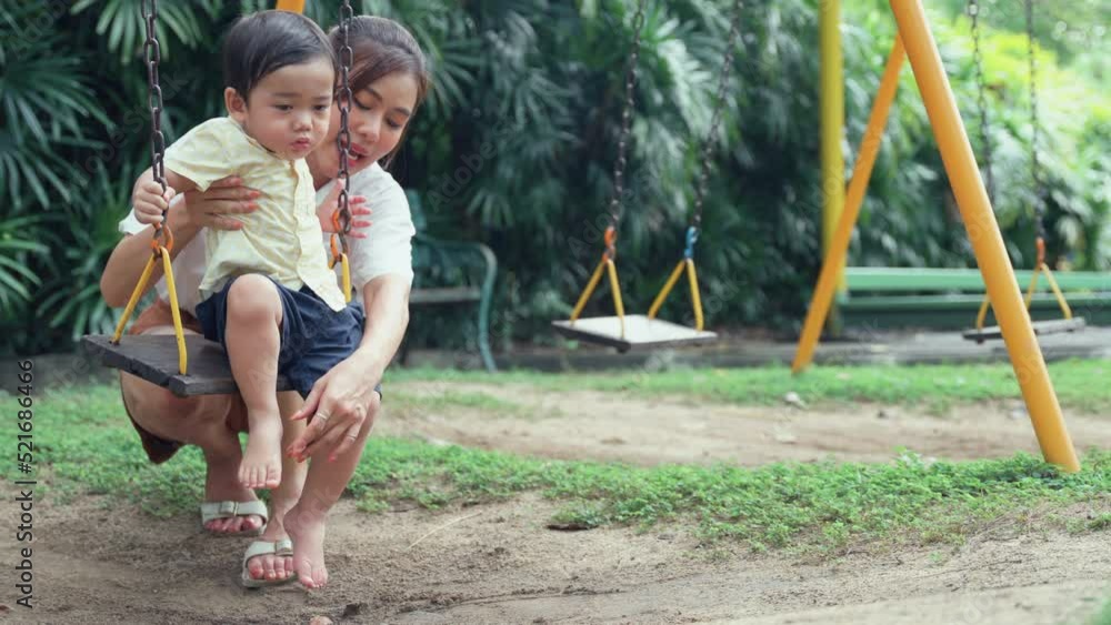 Asian young mother dusting off her lovely little son feet her son sitting on a swing in playground. Asian young mother dusting off her lovely little son feet her son sitting on a swing in playground.