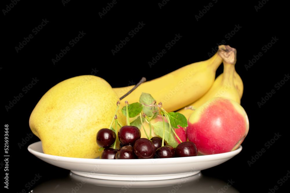 Several ripe sweet cherries, bananas, apples and a pear on a white ceramic plate, close-up, isolated on a black background.