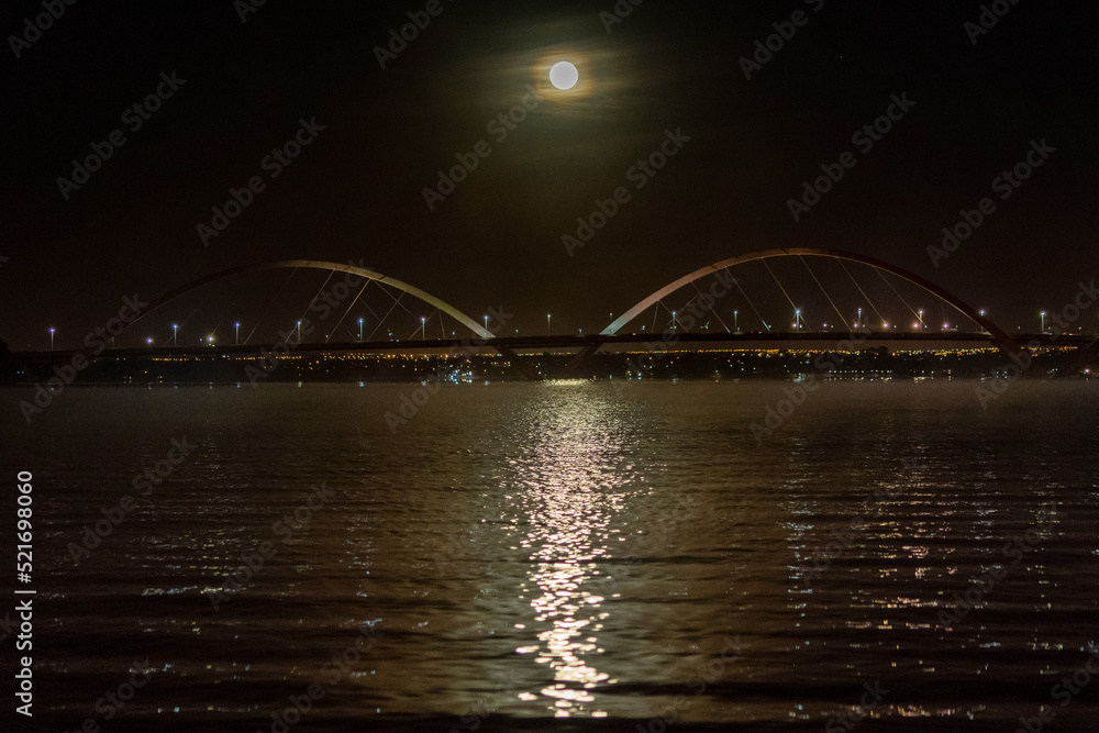 Naklejka premium The moon sunset over the JK Bridge in Brasilia, Brazil. Fog over Paranoa Lake. Supermoon rays reflecting in the water.